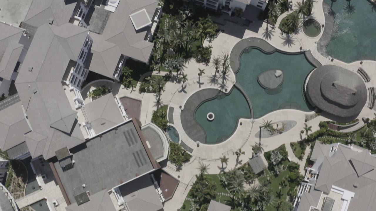 Overhead San Jose del Cabo resort view with the curved pool centered near the top, hotel roofs along the sides, palm planting, and no visible people.