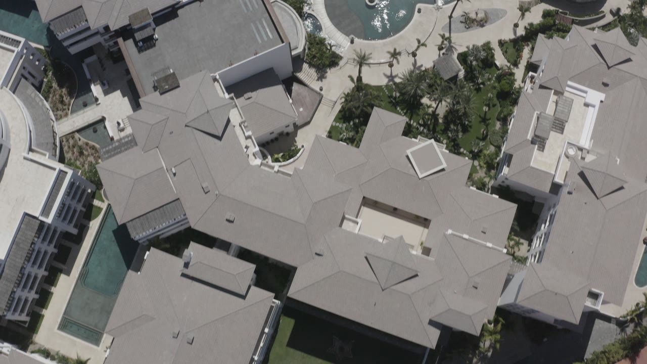 Overhead view of pale roofed buildings, pools, palms, and paved lanes.