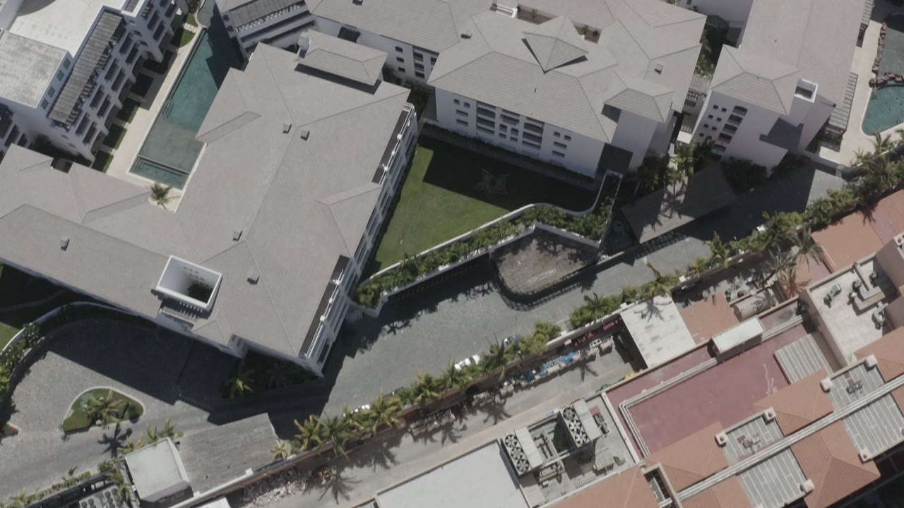 Top-down aerial frame of modern buildings and paved walkways in San Jose Del Cabo, Mexico, with red and gray rooftops, planted edges, and no visible people.