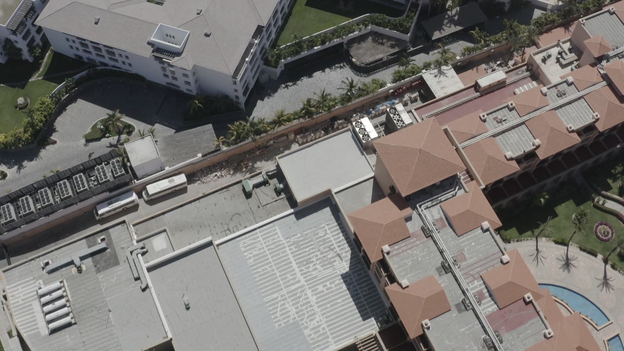 Aerial frame over San Jose Del Cabo, Mexico showing orange tile roofs, large flat gray rooftops, road edges, landscaped beds, and no visible people.