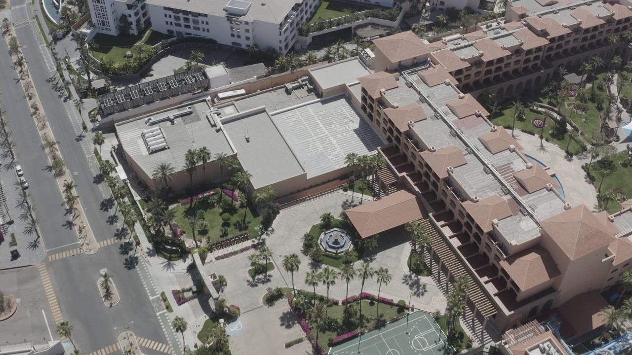 High aerial view over San Jose del Cabo, Mexico, with peach-roofed buildings, palm-lined streets, a sport court, landscaped courtyards, and no visible people.