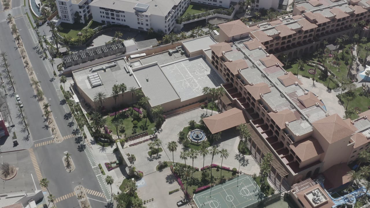 Top-down city block with large roofs, palms, courtyard planting, and sport court markings.