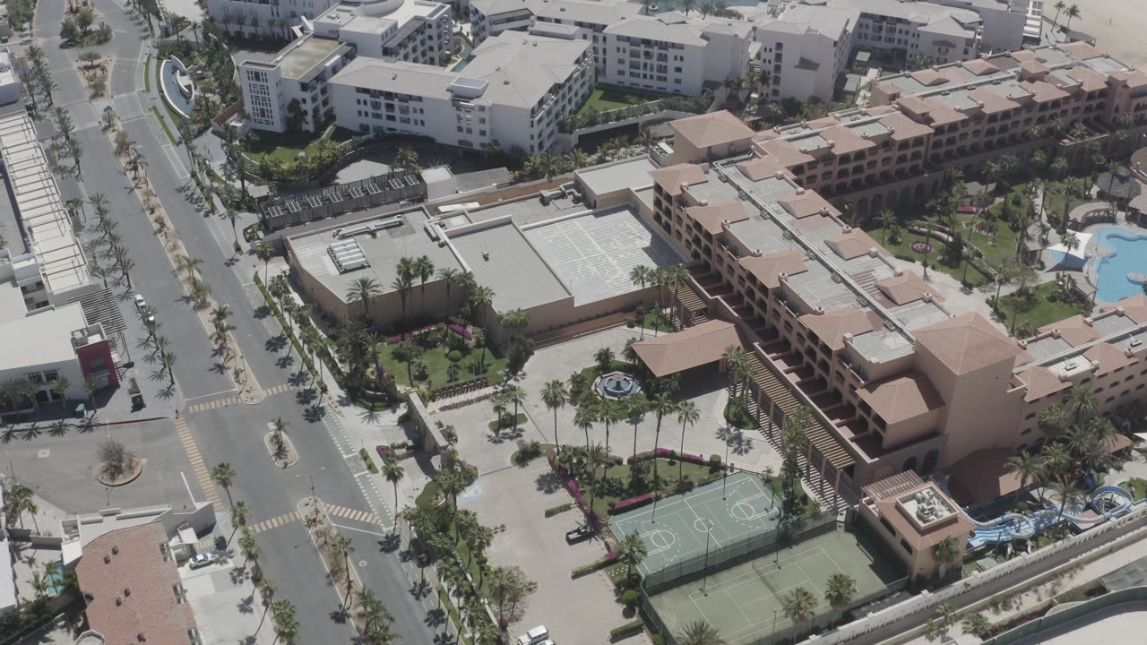 Aerial view of roads, resort buildings, pools, palms, and tennis courts near the coast.