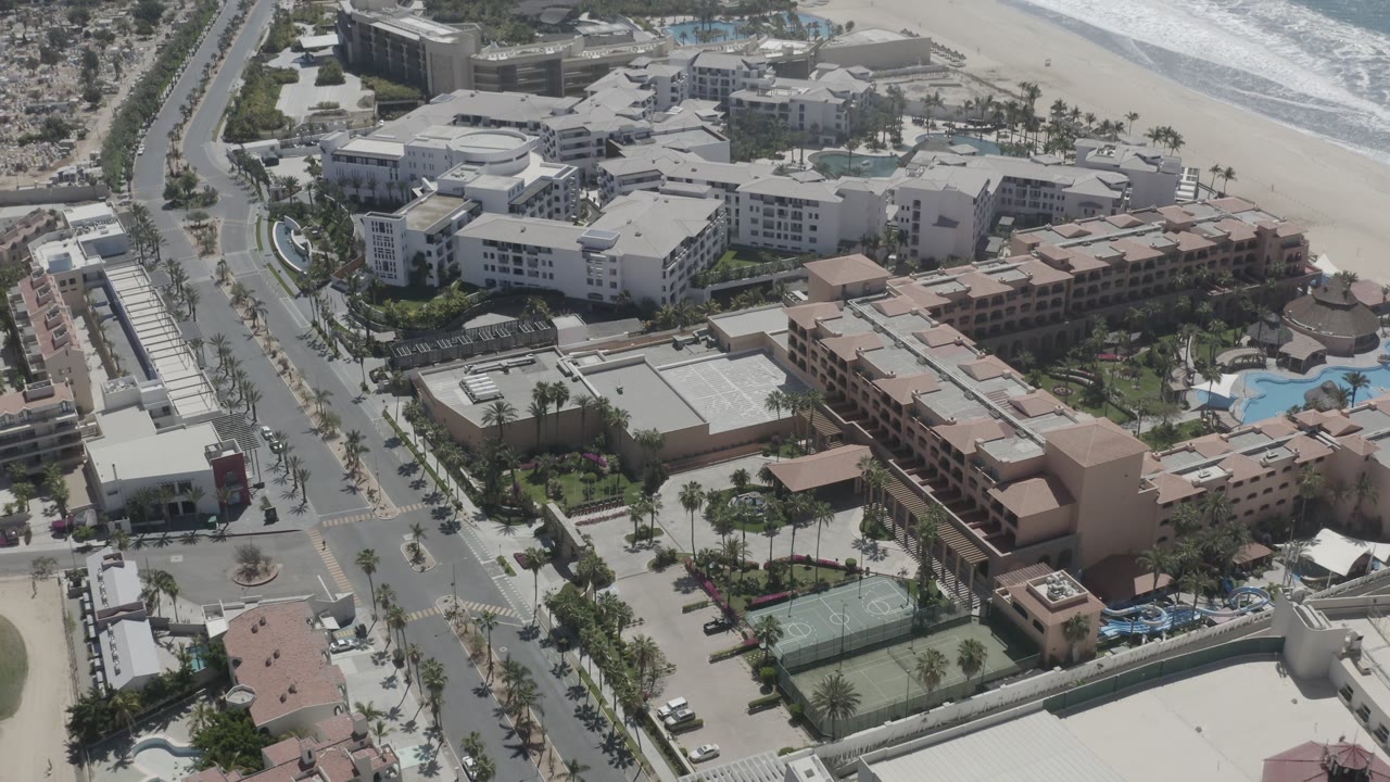 San Jose del Cabo aerial frame with the central resort block, nearby roads, parked cars, pools, tennis court, beach sand, and ocean water beyond, with no identifiable people visible.
