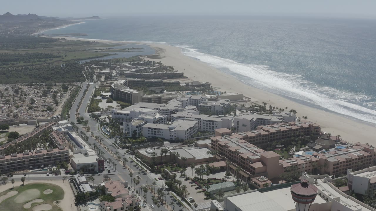 Aerial frame of San Jose del Cabo showing a beachfront district below, a long pale beach, white surf bands, blue-gray ocean, distant coastline, and no visible people.