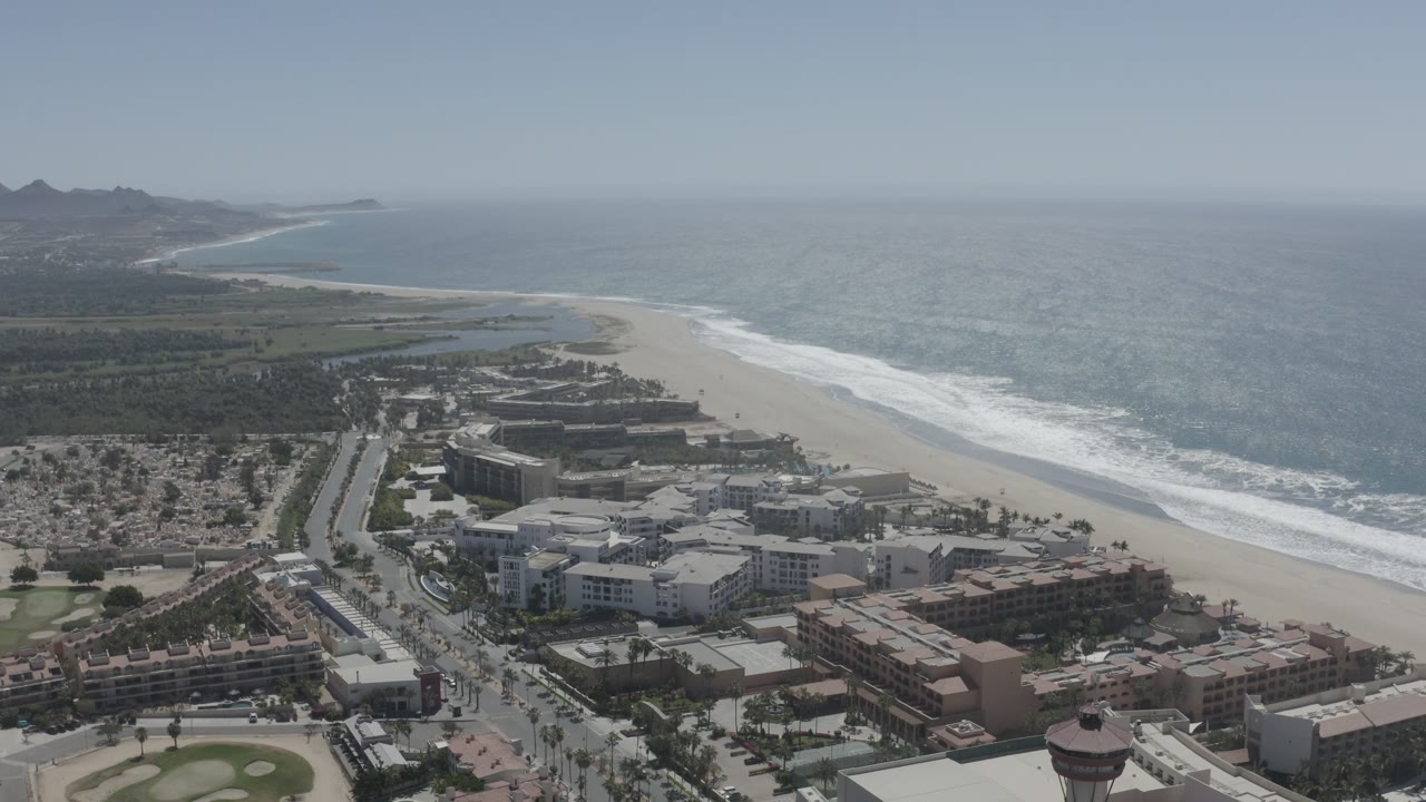 San Jose del Cabo coastline remains in a wide elevated view, with the beach curving past city blocks toward the hazy hills and no visible people.
