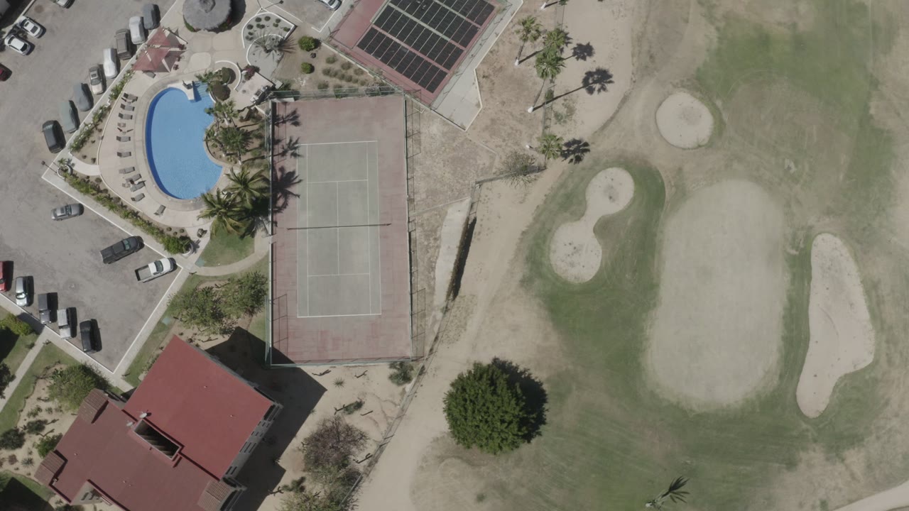 Top-down aerial frame in San Jose del Cabo, Mexico, showing a red-roofed building at lower left, a curved blue pool, tennis courts, parked cars, sand areas, and no visible people.