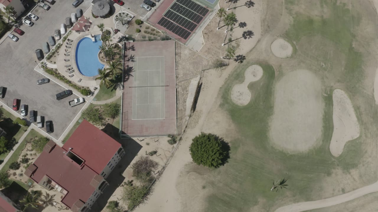 Aerial frame in San Jose del Cabo showing the blue pool near palms, the tennis court, red-roofed buildings, pale bunkers, and golf turf with no visible people.