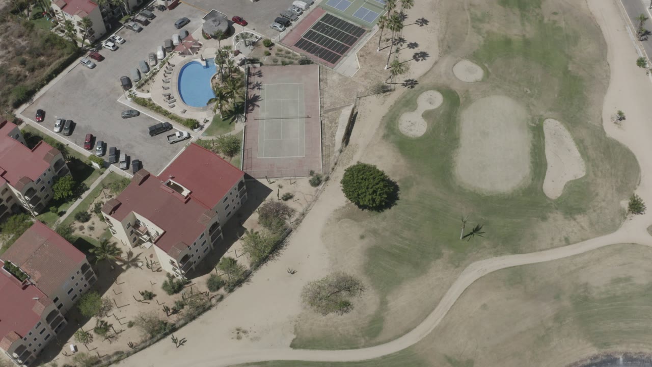 Aerial view in San Jose Del Cabo, Mexico, showing red-roof hotel buildings, a blue pool, tennis courts, parking, and golf greens with no visible people.