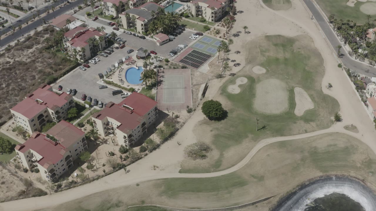 Aerial frame in San Jose del Cabo with the resort shifted slightly left, showing the pool courtyard, tennis courts, adjacent road, pale golf course grass, and no visible people.