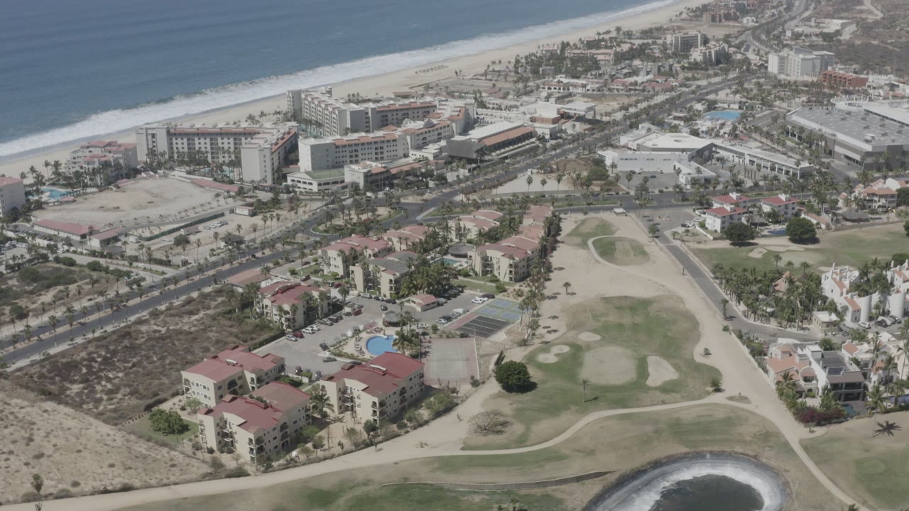 Aerial frame over San Jose del Cabo showing ocean surf, a wide beach, hotel-like buildings, red-roofed homes, roads, and green open space, with no individually discernible people.