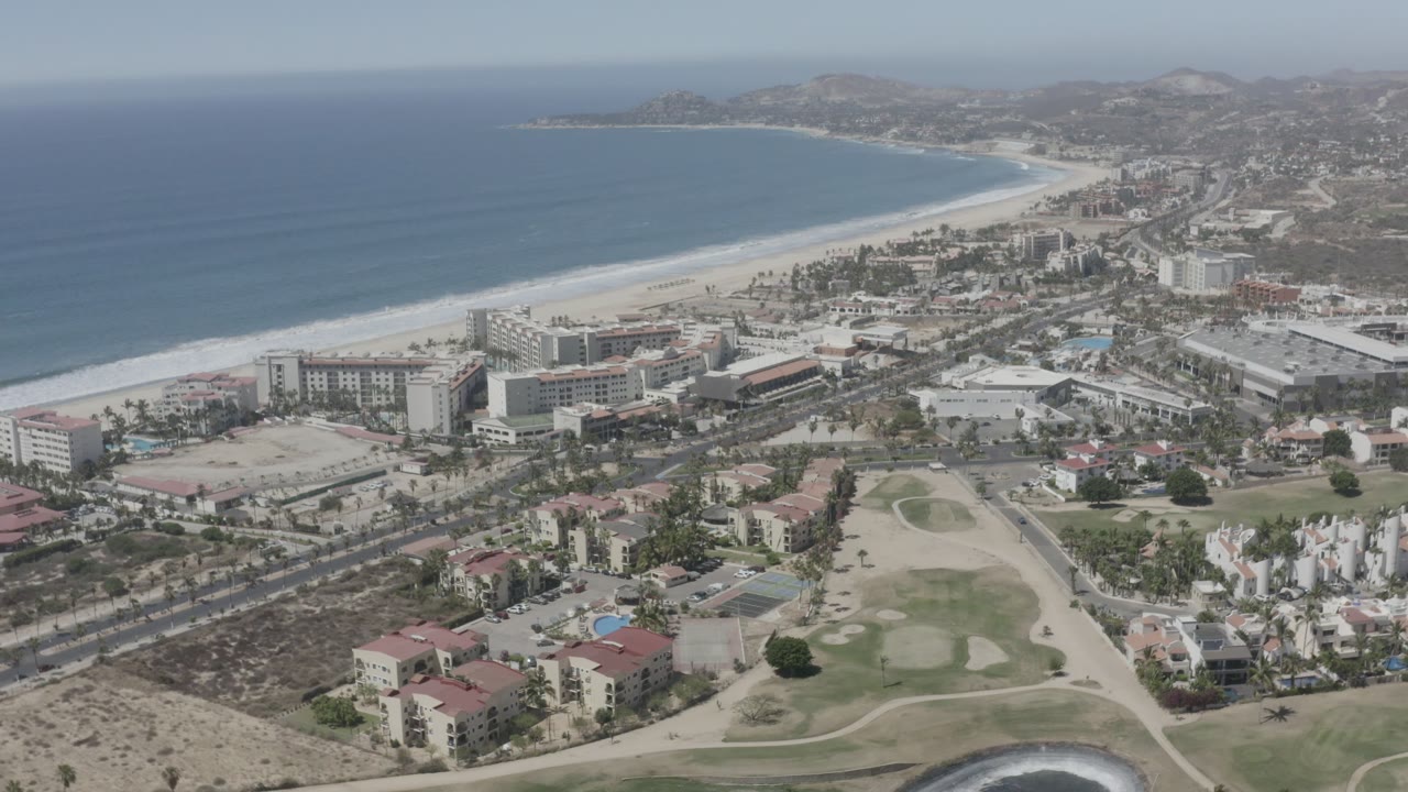 Wide San Jose del Cabo aerial showing the curved beach and ocean beside hotels, street grids, palm-lined areas, homes, and dry hills under hazy daylight, with no recognizable people visible.