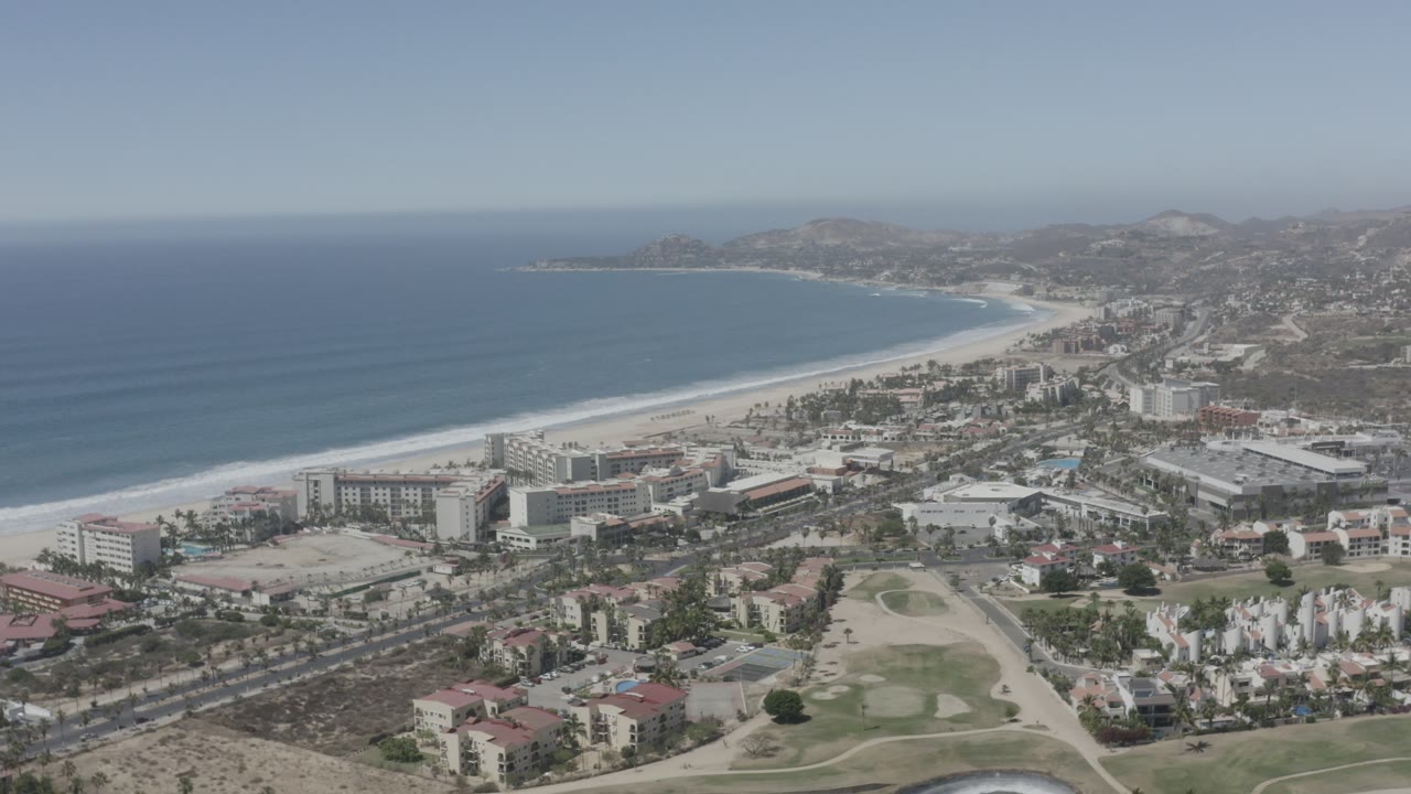 Wide aerial view of San Jose Del Cabo, Mexico, with calm blue water, a pale beach, resort blocks, nearby roads, scattered palms, and hillside development receding into haze.