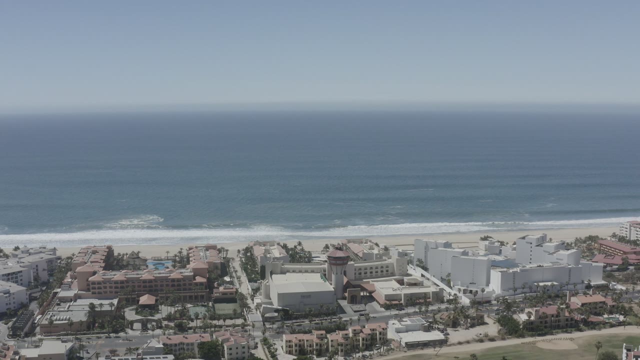 Aerial-looking view of San Jose del Cabo beachfront buildings below a sandy beach and blue ocean, with no visible people.