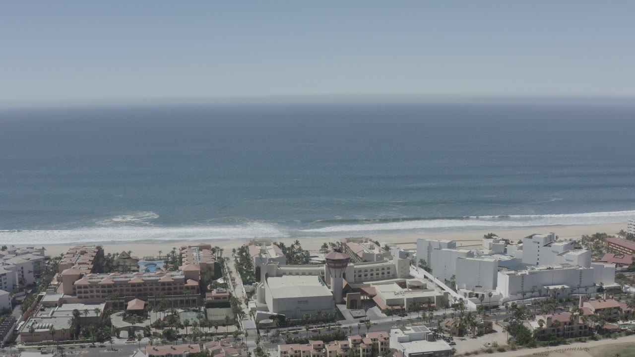 Aerial frame over San Jose del Cabo, Mexico with blue ocean beyond white surf, a sandy beach, and beachfront buildings in the lower frame with no visible people.