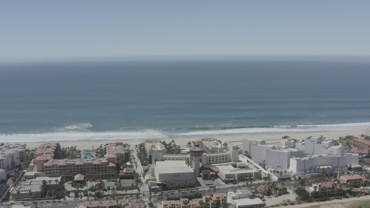 San Jose del Cabo aerial view with the horizon high in frame, rolling surf along the beach, and white and terracotta-roofed coastal buildings below with no visible people.
