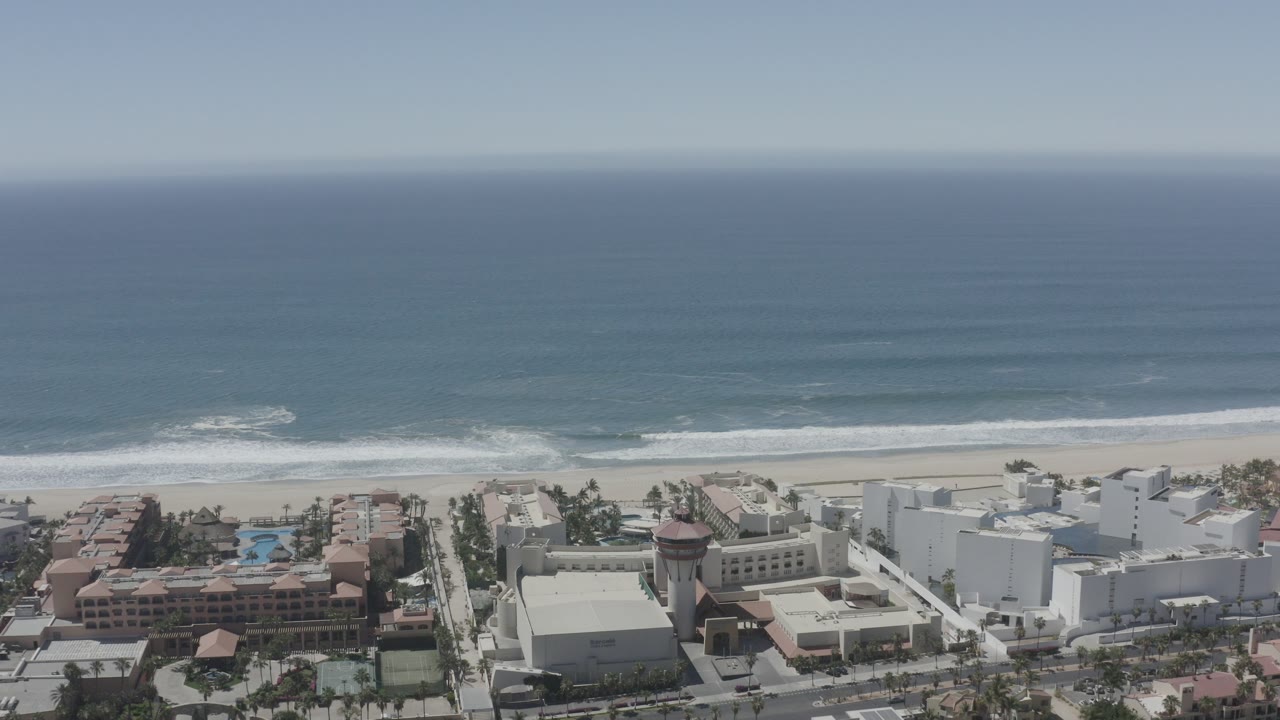 High aerial frame over San Jose del Cabo resort buildings, sandy beach, blue ocean bands, and no visible people at the start of the clip.