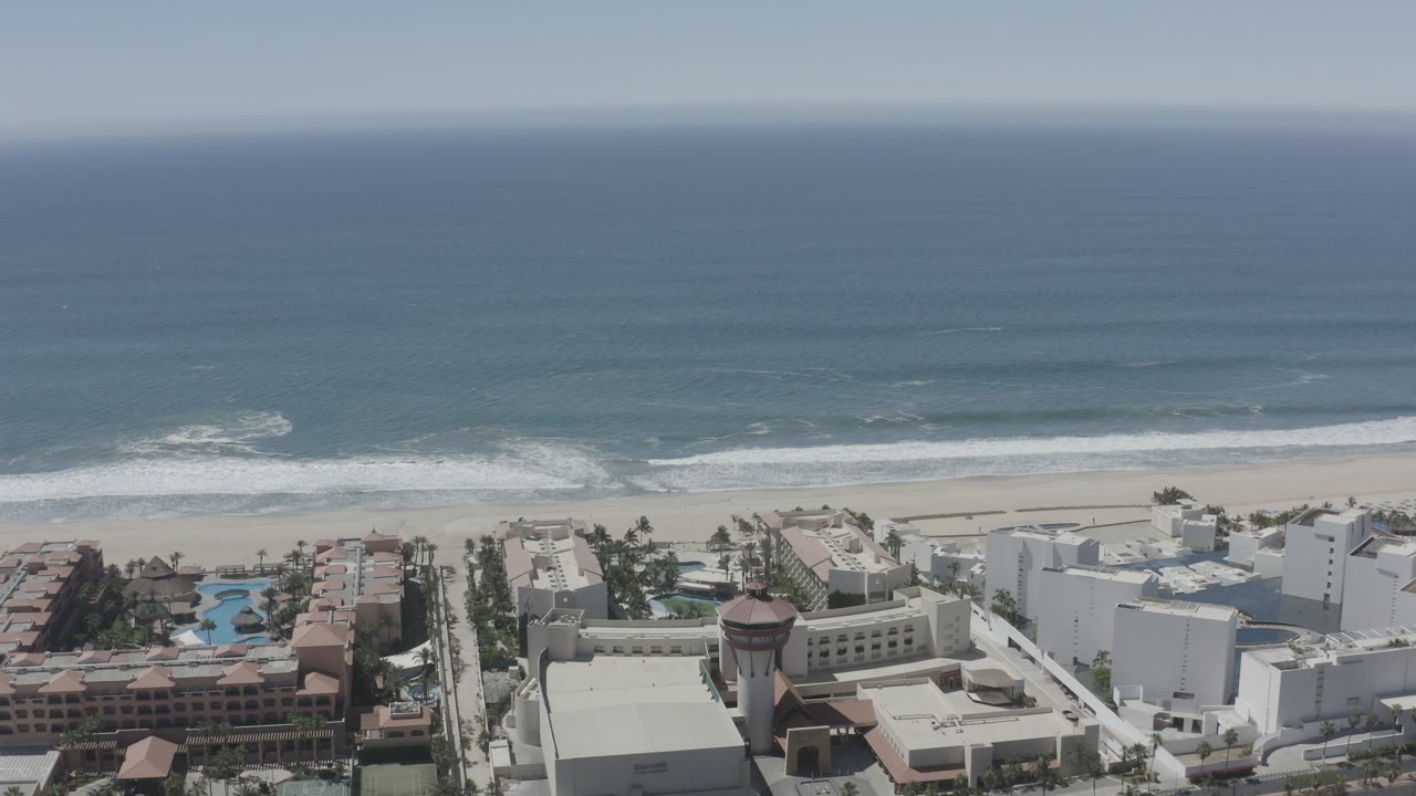 A high aerial frame over beachfront apartments and resort buildings in San Jose Del Cabo, Mexico, with pools below, pale sand beyond, rolling surf, blue ocean, and no visible people.