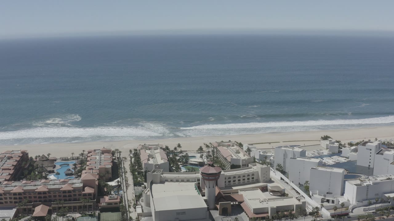 The aerial view holds on San Jose Del Cabo beachfront buildings, with white rooftops, terracotta apartment blocks, pool areas, breaking waves, and no visible people.