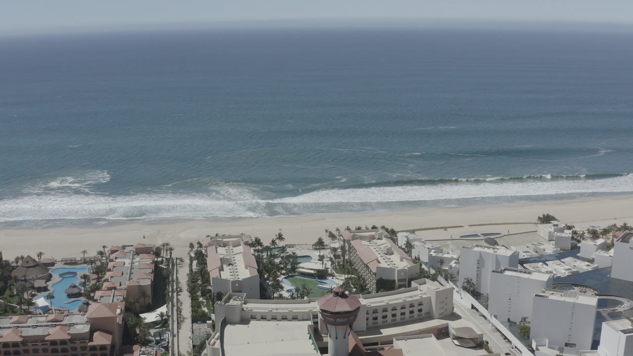 Opening aerial frame over San Jose del Cabo with blue ocean, white surf, pale beach, pools, palms, and beachfront buildings, no visible people.