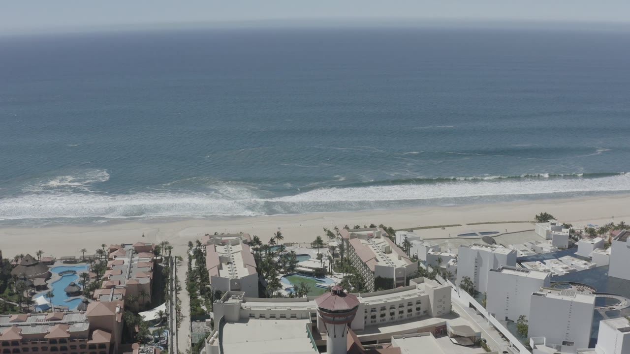 San Jose del Cabo aerial view with a broad surf band nearing the beach and hotel-style buildings and blue pools in the lower frame, no visible people.
