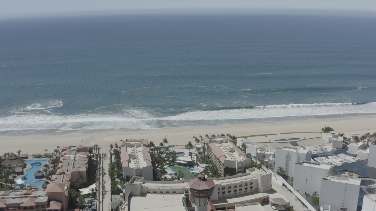 High drone view of San Jose del Cabo shoreline with darker blue water offshore, white foam at the beach, palms, pools, and white oceanfront buildings, no visible people.