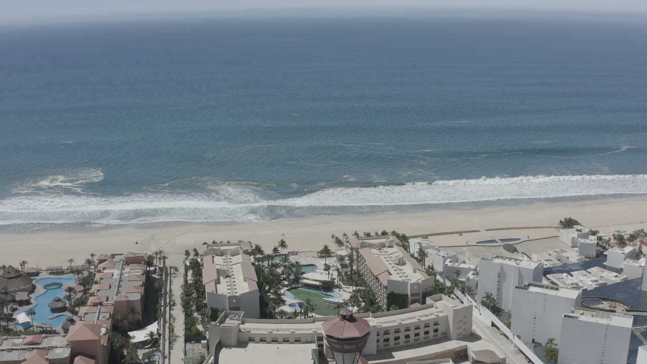 Aerial beach scene with surf, sand, pools, and resort-style rooftops.