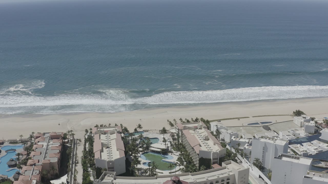 Aerial coastline with blue ocean, sandy beach, beachfront buildings, pools, and palm trees.