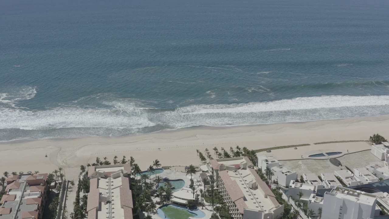 Aerial view of blue ocean surf, sandy beach, and coastal buildings.
