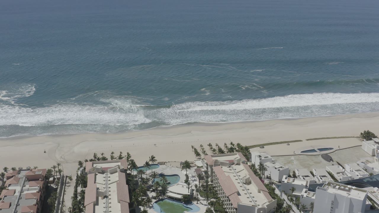 Aerial frame in San Jose del Cabo, Mexico, with a white wave line curling toward the sand and coastal buildings, pools, and palms along the lower edge, with no visible people.