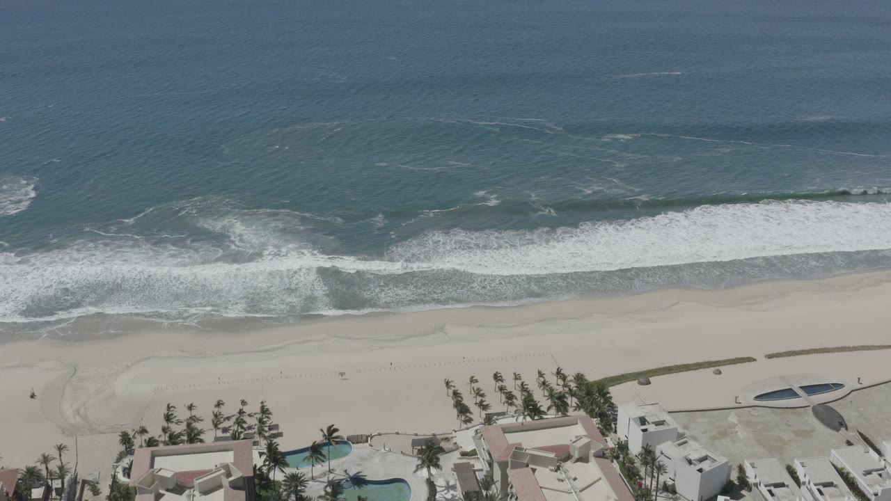 Aerial view of surf breaking onto a sandy beach beside palms and resort buildings.
