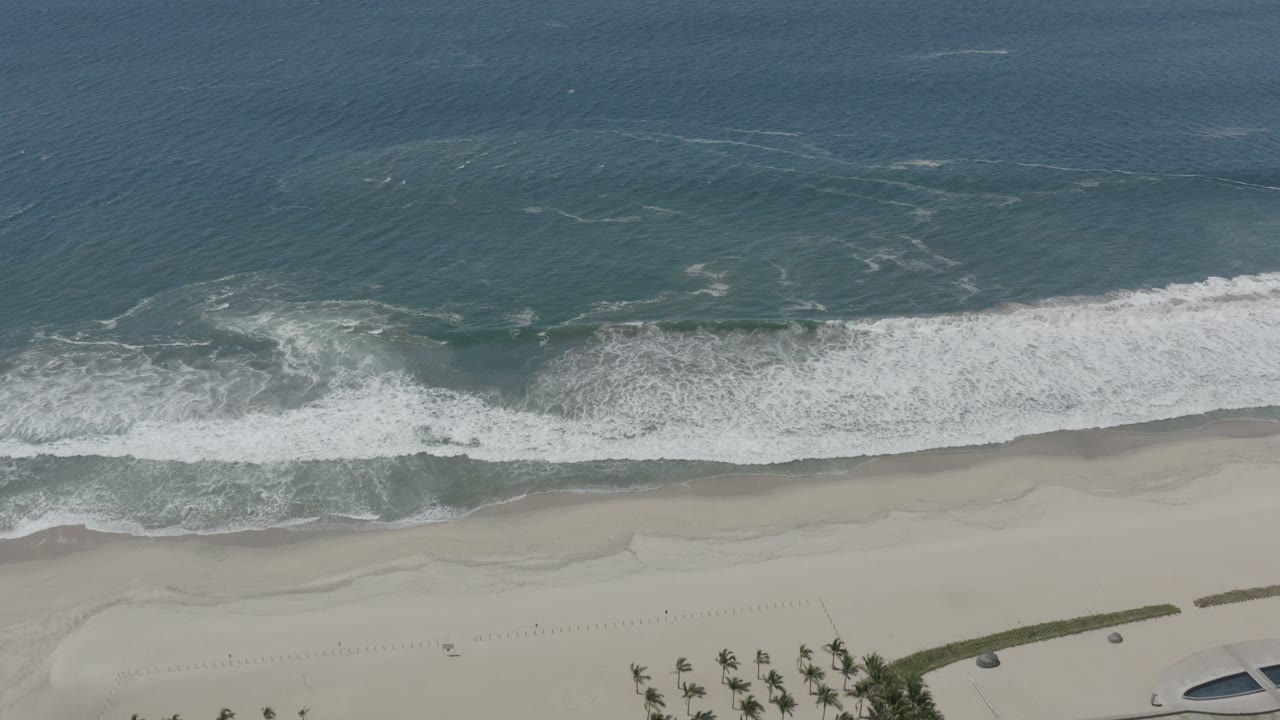 Overhead drone view of blue-green water and white surf reaching a pale beach in San Jose del Cabo, with palms near the lower edge and no visible people.