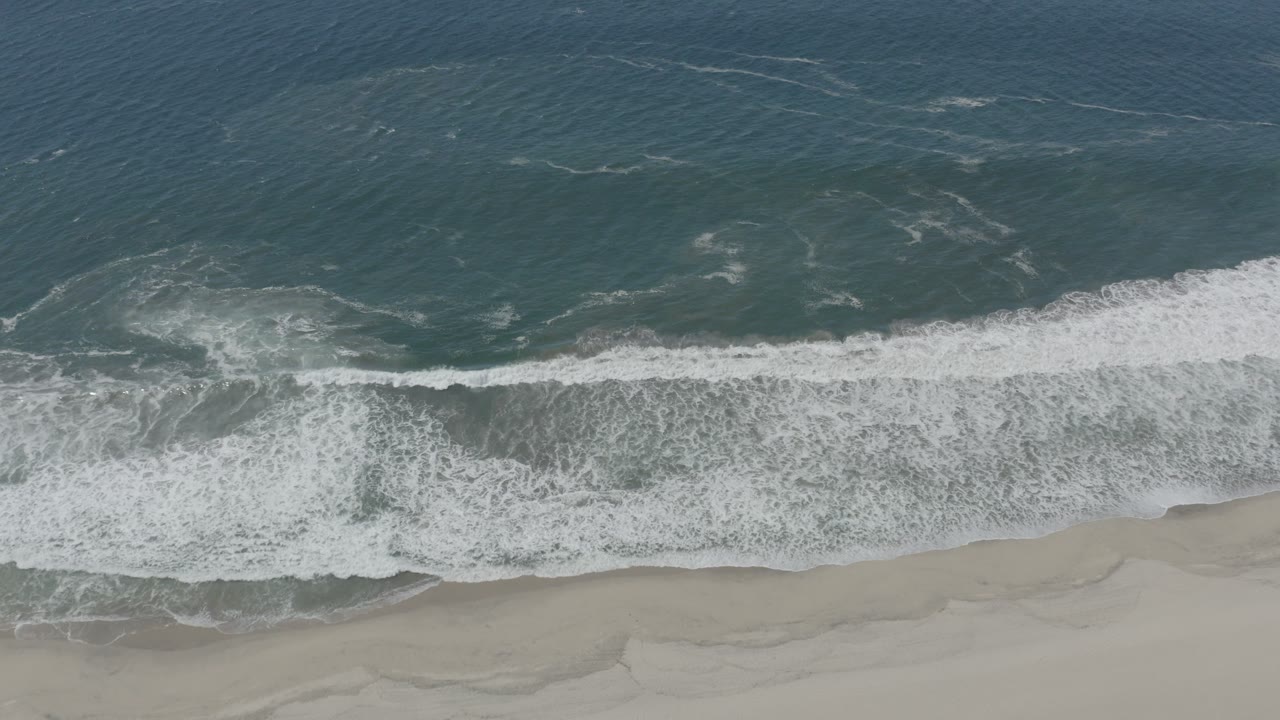 Overhead view in San Jose del Cabo, Mexico, with dark blue-green ocean above, white surf breaking across the middle, pale sand below, and no visible people.