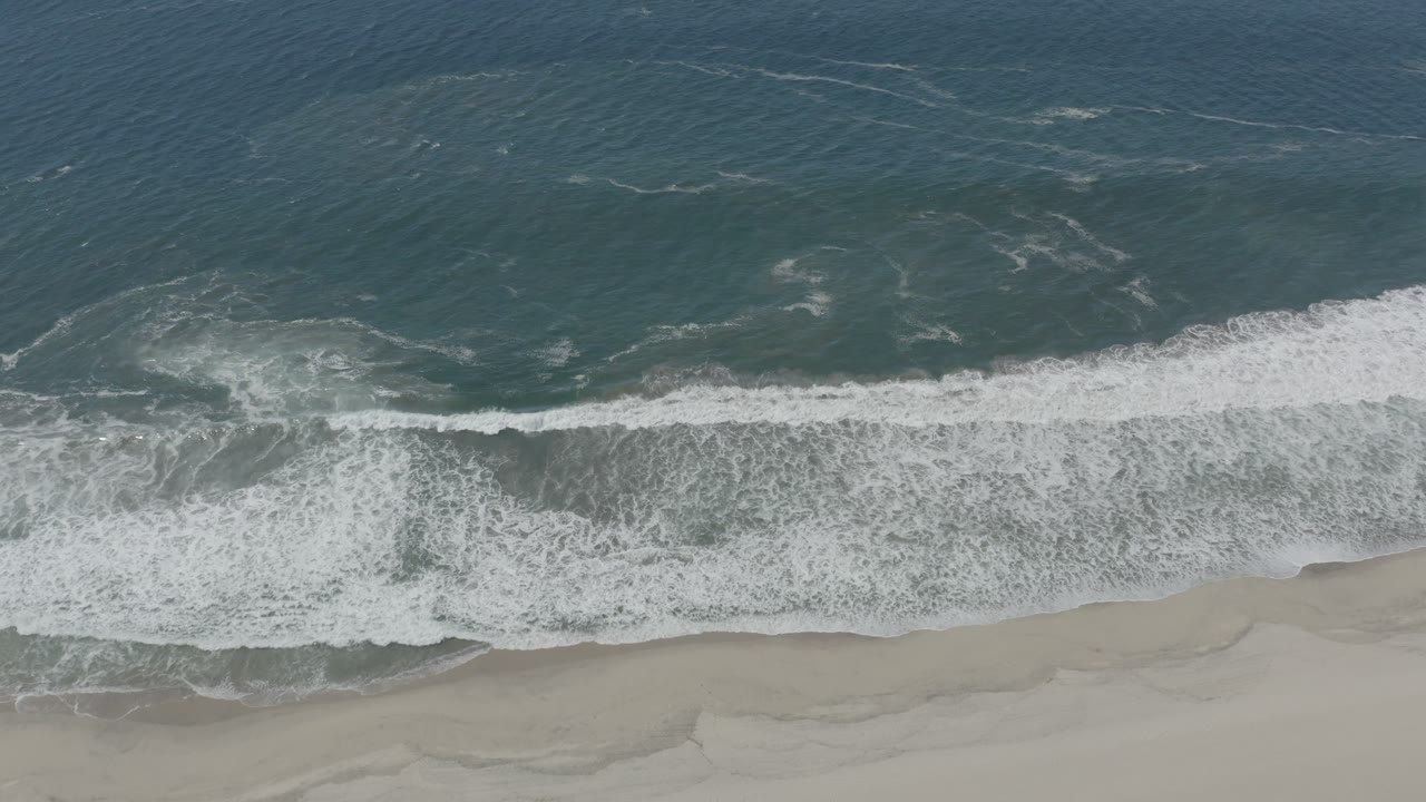 Aerial frame from San Jose del Cabo, Mexico, showing white foam spreading diagonally over the shoreline with darker ocean water filling the upper half and no visible people.