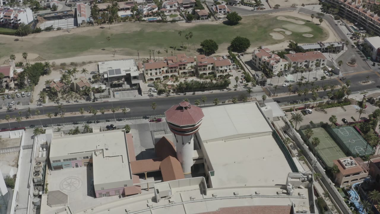 Opening aerial frame over San Jose del Cabo showing a red-roofed tower, nearby flat-roofed buildings, a road, tennis courts, housing, golf fairways, and no visible people.