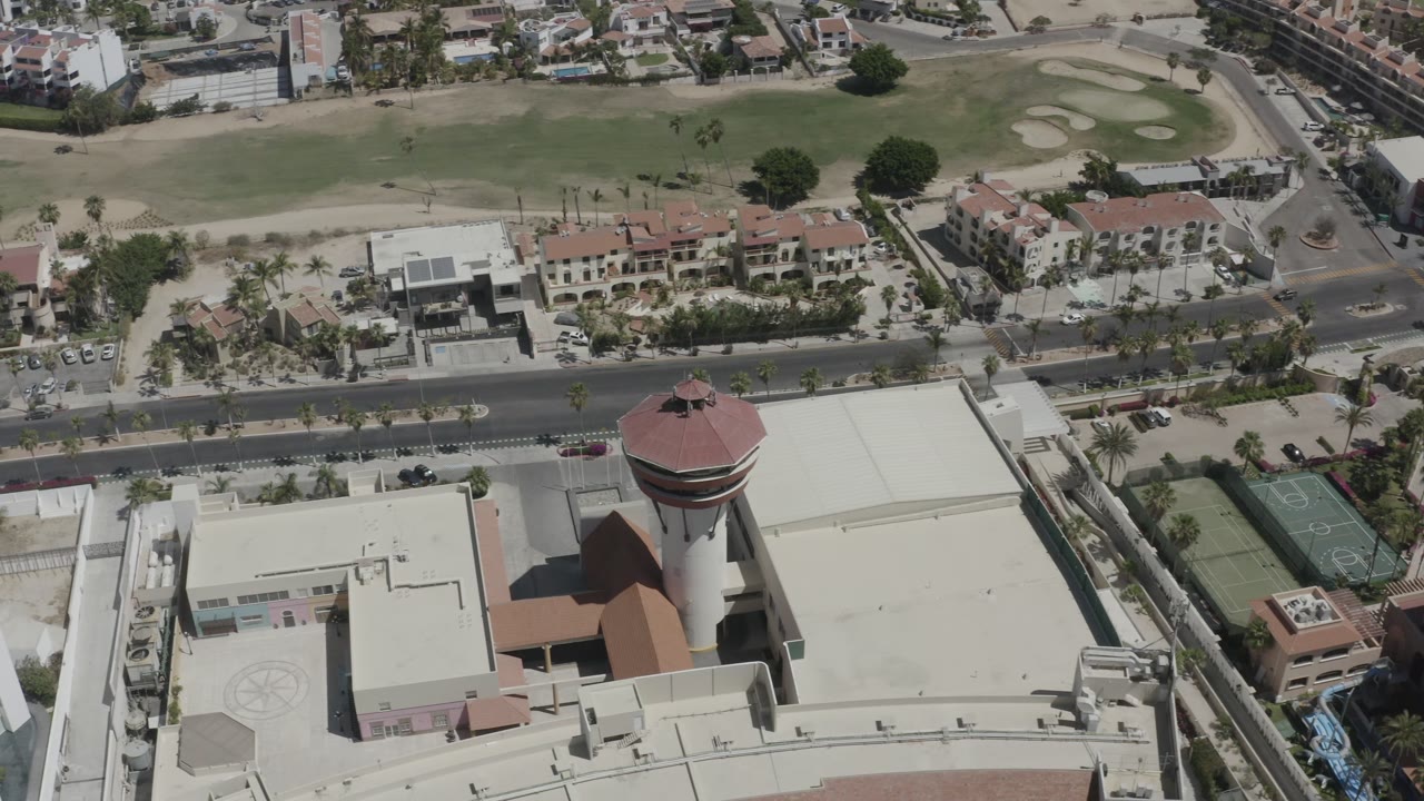 Aerial frame in San Jose del Cabo with the red-roofed tower centered lower right, apartments across the road, golf course grass and sand traps in the background, and no visible people.