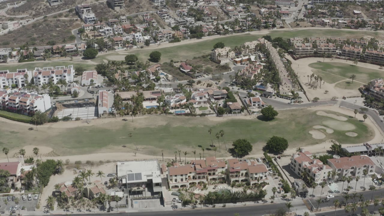 Aerial frame over San Jose del Cabo showing green fairway, sand bunkers, white residential blocks, terracotta roofs, dry hillside housing, and no visible people.