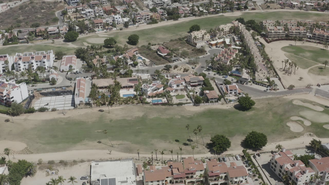High aerial frame over San Jose del Cabo with a golf fairway in front, low-rise homes and roads beyond it, and no visible people.
