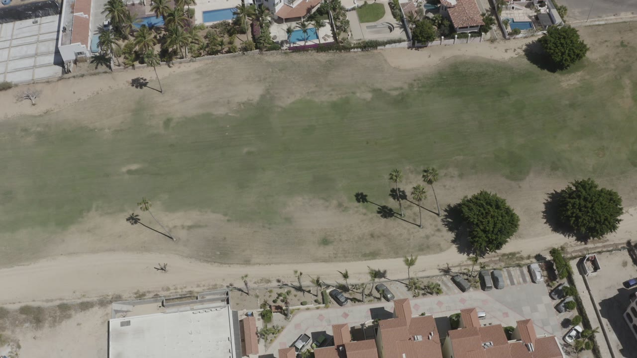 Overhead view of shallow green water, sandy shore, villas, pools, palms, and parked cars.