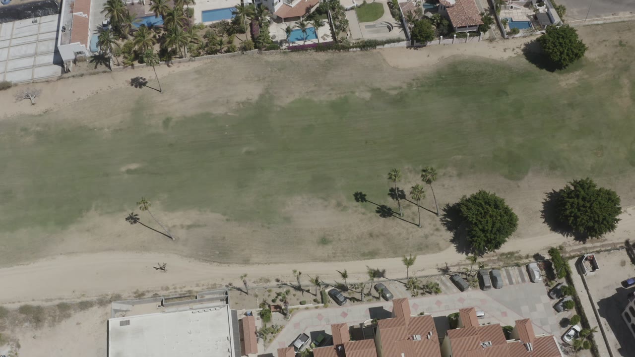 Drone-style overhead frame in San Jose del Cabo with a wider band of shallow water, a pale beach path, villas with pools near the top, palm clusters, parked cars, and no visible people.
