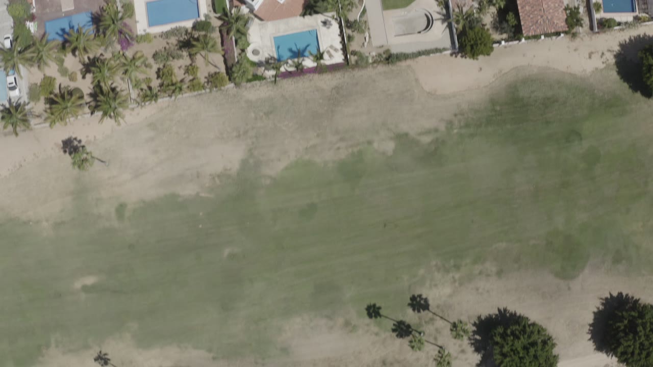 A high overhead frame in San Jose del Cabo, Mexico, with tan-roof homes, blue pools, palms, sand, and green coastal water, with no visible people.