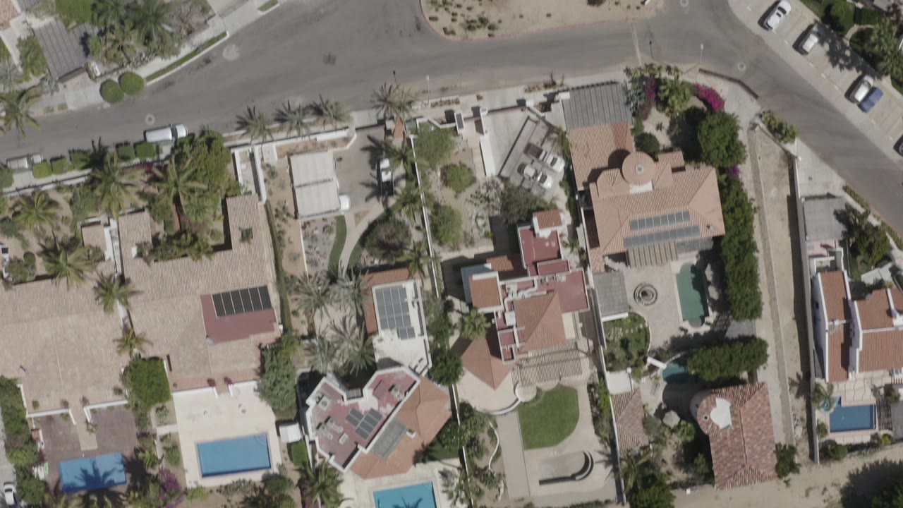 Top-down aerial frame over San Jose del Cabo homes with walled yards, blue swimming pools, palm trees, parked cars, and no visible people.