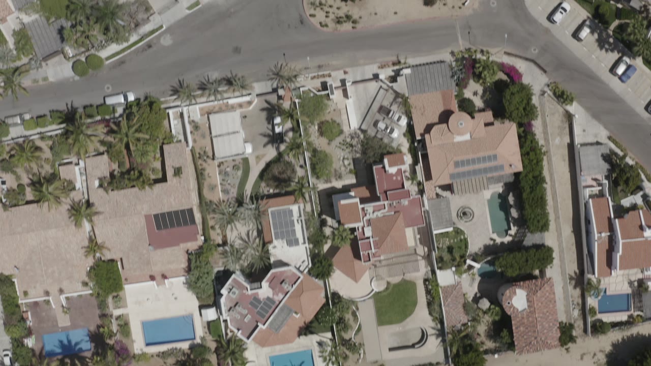 Overhead San Jose del Cabo neighborhood view with tan rooftops, rectangular and curved pools, landscaped yards, a road junction, and no visible people.