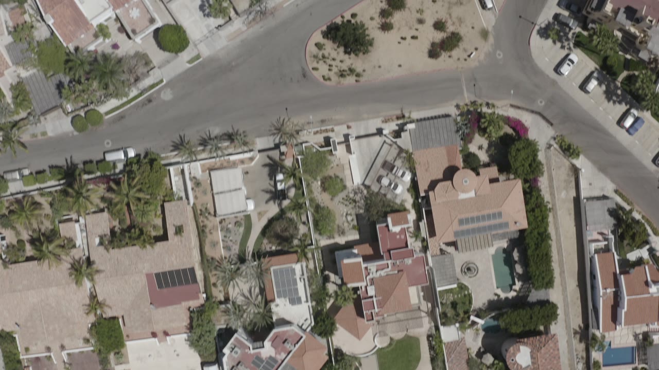 Downward aerial view of villas, palms, pools, and parked cars along streets.