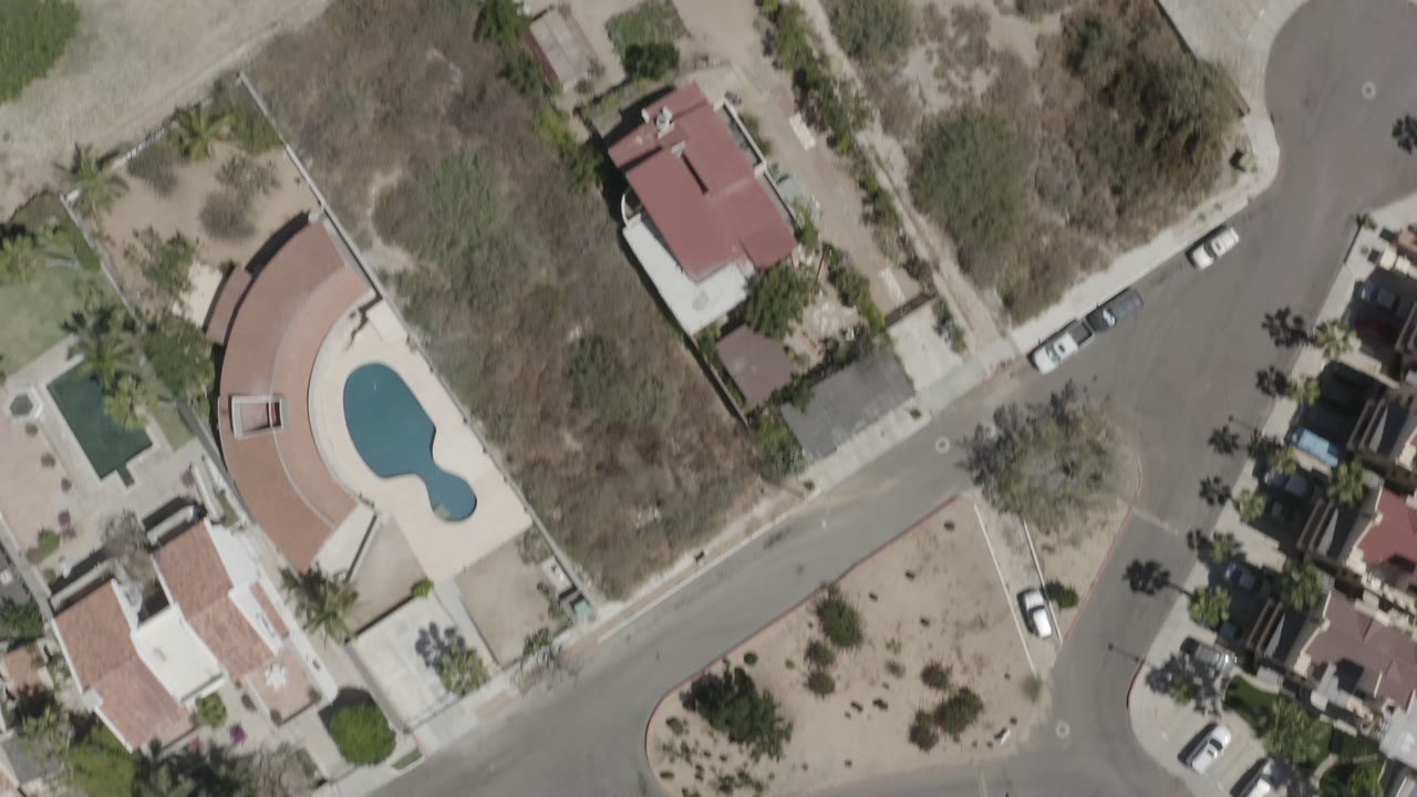 Aerial frame in San Jose del Cabo with a red-roof home, dry open lots, intersecting residential roads, parked cars, and tree shadows, with no visible people.