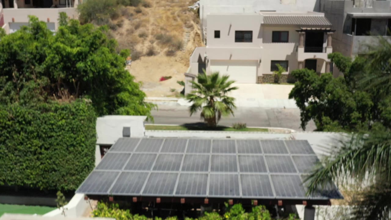 Aerial frame in San Jose del Cabo with the solar-panel roof still prominent below, a palm and trees near the road, pale modern houses uphill, dry brush, and no visible people.