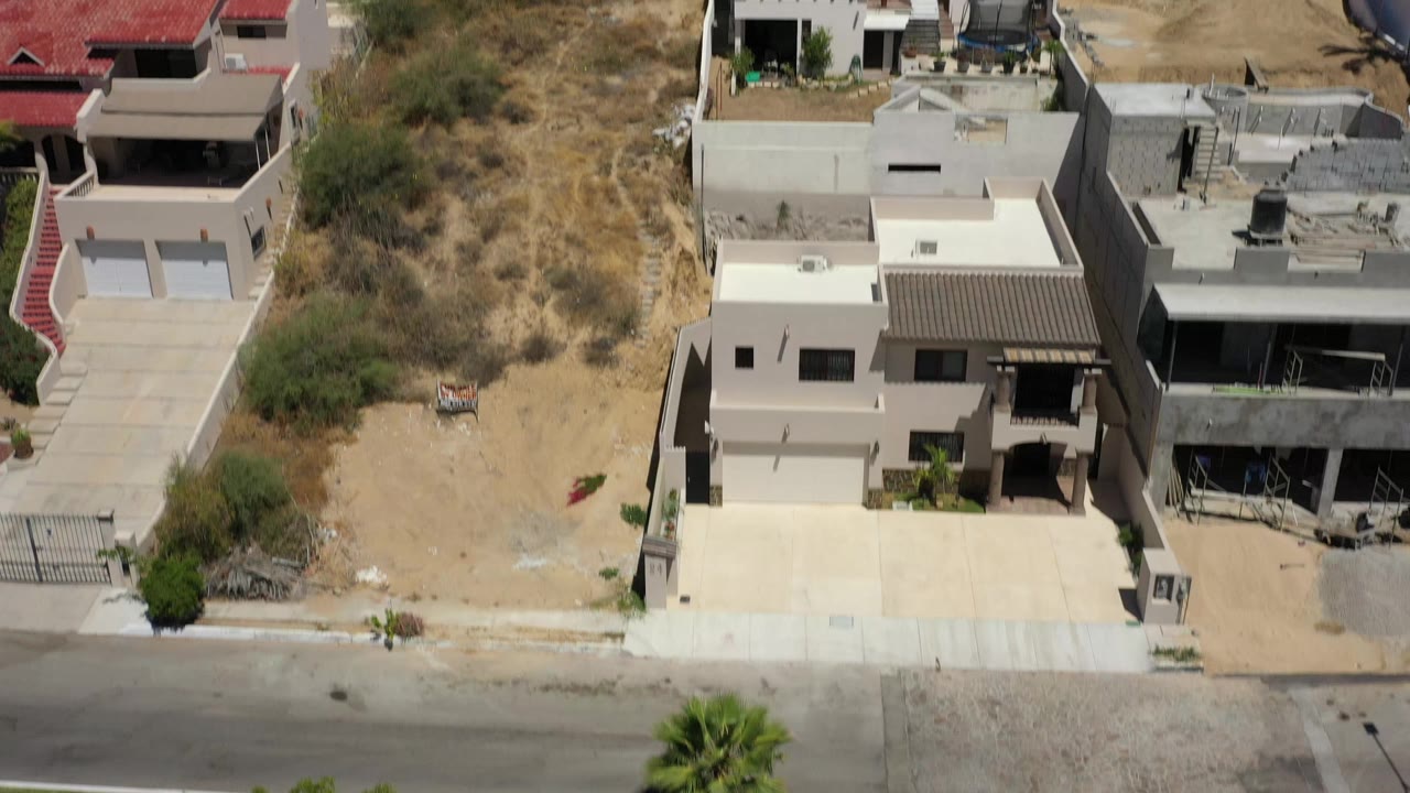 High-angle San Jose del Cabo view showing adjoining homes, walled property lines, a paved street, tan hillside vegetation, and no visible people.