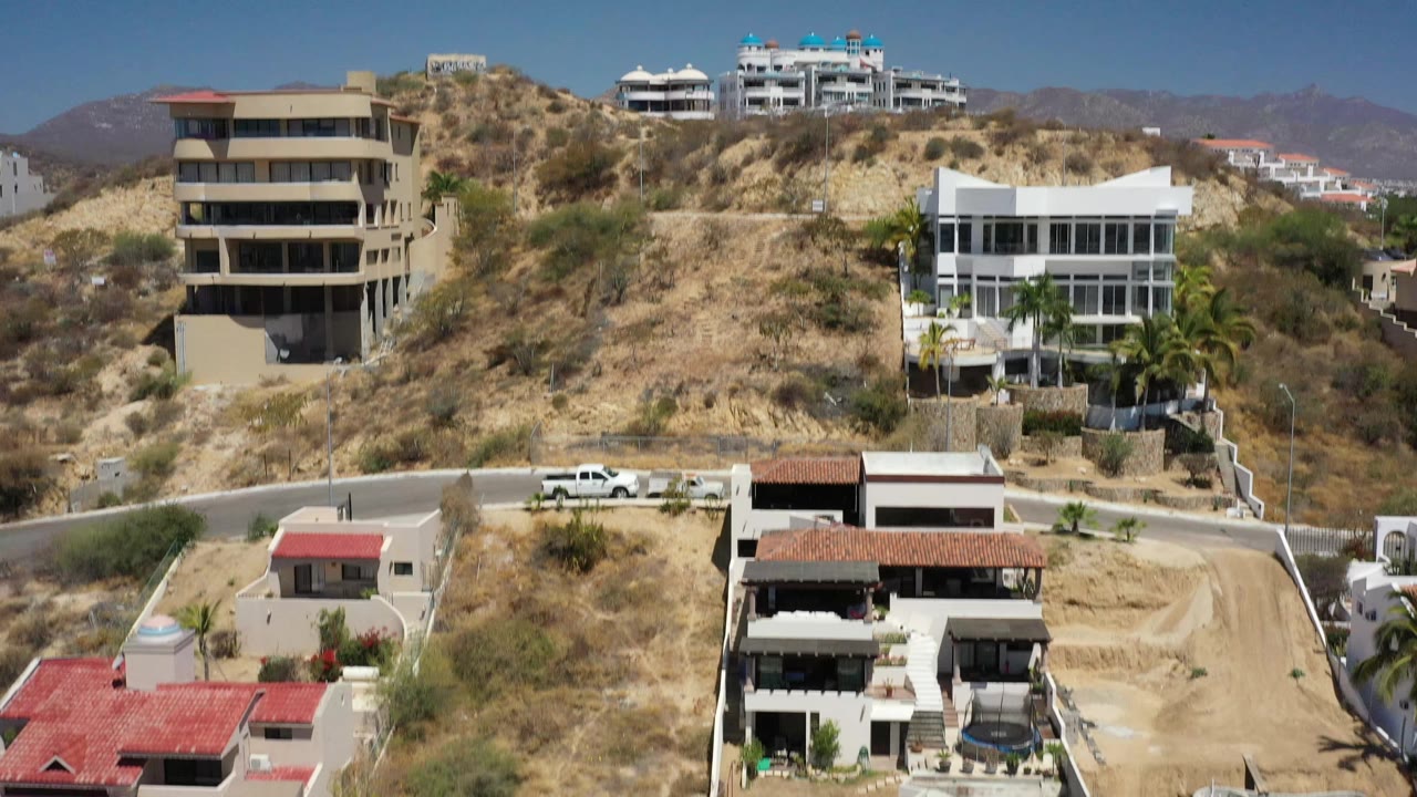 Elevated 4K view of San Jose del Cabo hillside residences, showing a beige tower-like building, a white modern house, dry scrub, mountains, and no visible people.