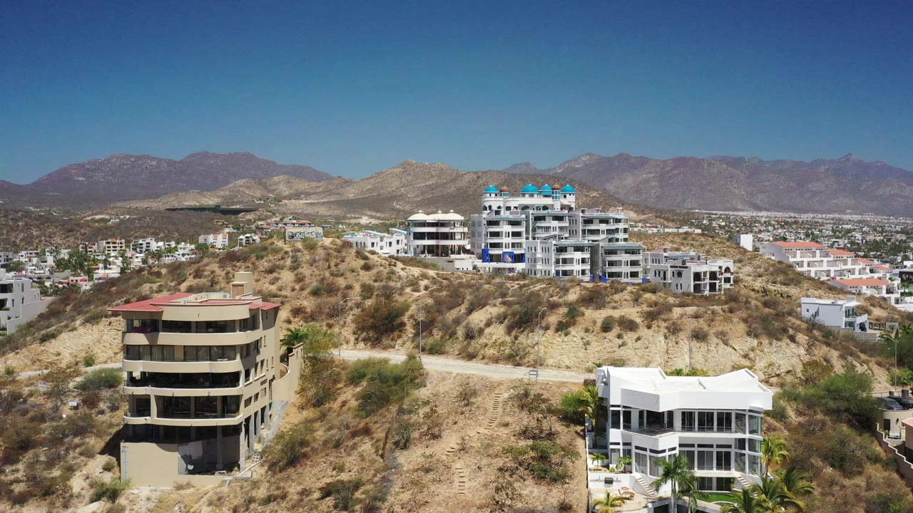 High aerial frame in San Jose del Cabo, Mexico, with a beige building in the lower left, white hillside residences near center, distant neighborhoods, mountains, and no visible people.