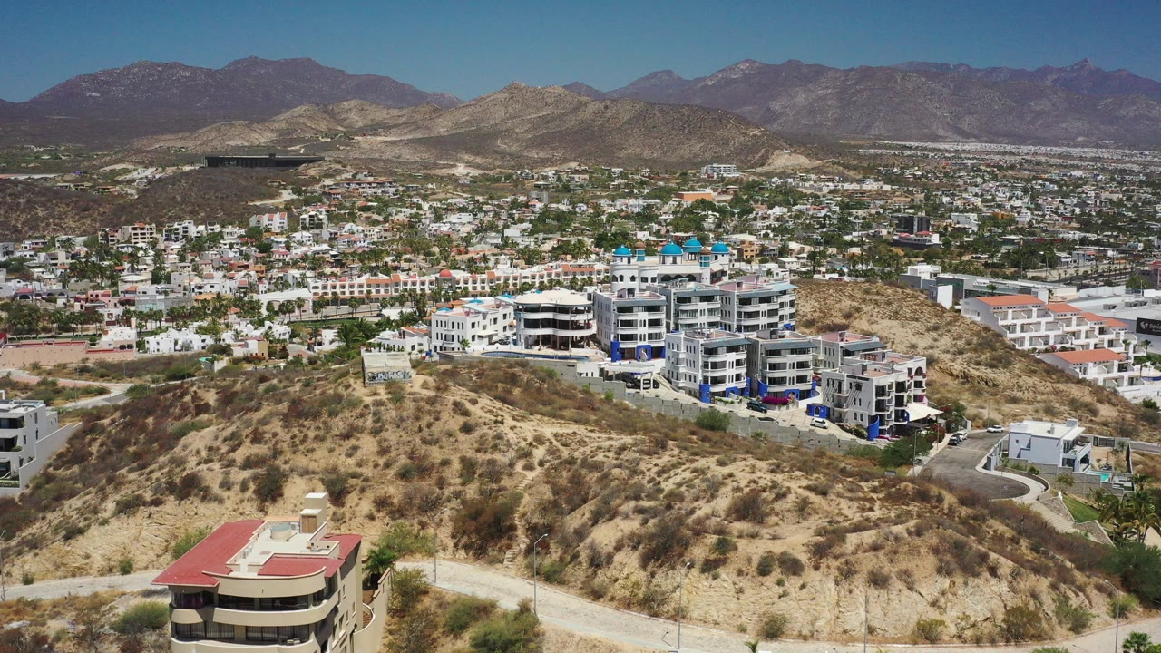 Opening aerial frame over San Jose del Cabo, Mexico, showing scrub-covered hills, white residential buildings, a blue pool area, distant city blocks, mountains, and no visible people.